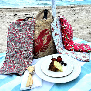 Colorful adult bibs on a  beach picnic setup with lobster-themed bag, blanket, and food on the sand.