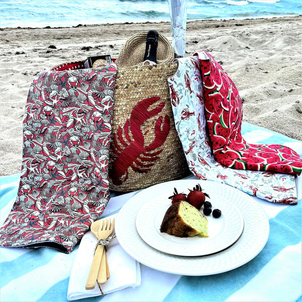 Colorful adult bibs on a  beach picnic setup with lobster-themed bag, blanket, and food on the sand.